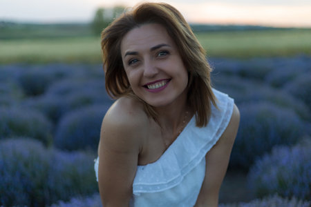 Beautiful and pretty young woman in a white dress relaxes and enjoys a walk in the lavender field at sunset, toned. Selective focus with blurred background.の写真素材