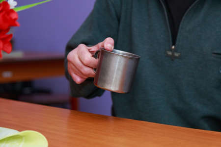Homeless man warms himself up in a shelter with a metal mug of hot tea.の写真素材