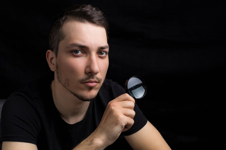Young handsome man and a magnifying glass on a black background in low key. Search symbol....