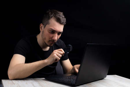 Young handsome man looks through a magnifying glass at the laptop screen on a black background in a low key. Search symbol. Investigation conceptの写真素材