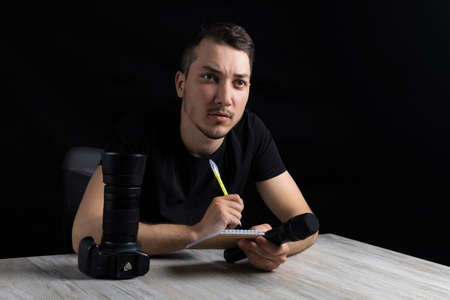 Young handsome man in black clothes with camera, microphone, notebook and pen, on black background...