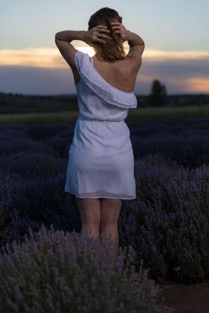 Beautiful and pretty young woman in a white dress relaxes and enjoys a walk in the lavender field at sunset, toned. Selective focus with blurred background.の写真素材