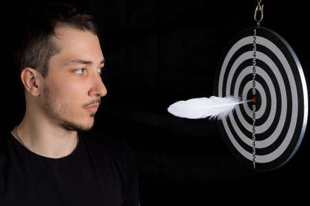 Young handsome man and dart board with a quill pen stuck in the center of the target on a black background in a low key. Writing author goal achievement conceptの写真素材