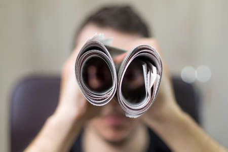 Man holding two twisted roll newspaper. Metaphor or allegory with binoculars. Selective focus on newspapers. Truth search conceptの写真素材
