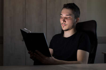 Young handsome man or an adult guy in the dark reads a glowing book. The concept of teaching is light, not teaching is darkness.の写真素材