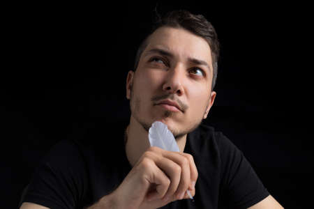 A young handsome man in black clothes with a goose feather looks pensively and romantically up, close-up, on a black background in a low key. Copyspace for textの写真素材