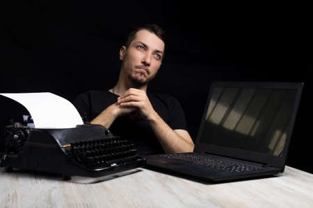 Young handsome man midway between a typewriter and laptop on a black background in low key. Creativity symbol. The concept of the past and the future. Time and changeの写真素材