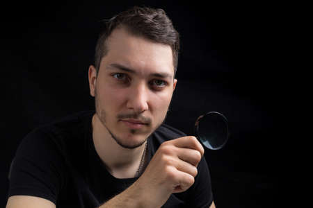 Young handsome man and a magnifying glass on a black background in low key. Search symbol. Investigation conceptの写真素材