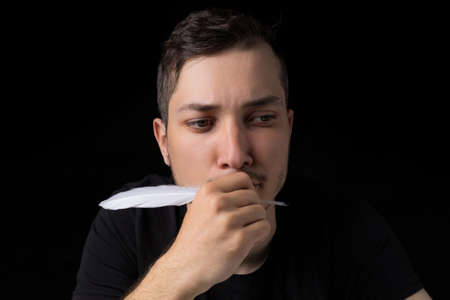 A young handsome man in black clothes with a goose feather looks pensively and romantically up, close-up, on a black background in a low key. Copyspace for textの写真素材