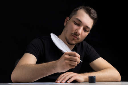 Young shy and smiling handsome man in black clothes dips a quill pen into an inkwell on a black...