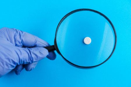 Classic medical pill with risk, doctor's hand in gloves and magnifying glass in blurred foreground. Traditional blue backgroundの写真素材