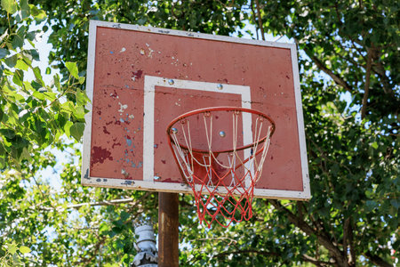Weathered outdoor basketball hoop with faded red backboard amidst green trees.