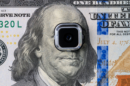 Surveillance camera lens overlaying benjamin franklin on us dollar bill.