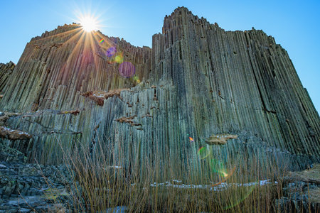 Sunlit basalt columns with grass in foreground under clear sky.