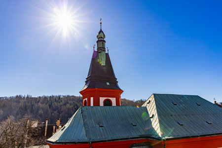 Sunlit church with red tower and green roof under clear blue sky in forested landscape.の写真素材