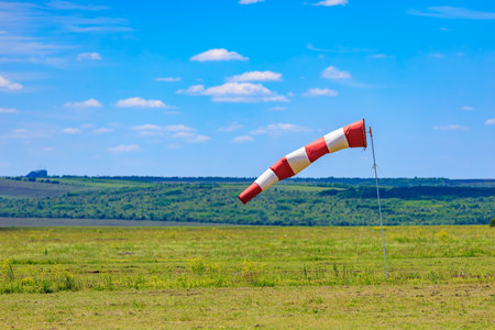 Red and white windsock over green meadow with blue sky and clouds.