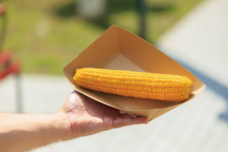 Hand holding grilled corn in cardboard tray outdoors.