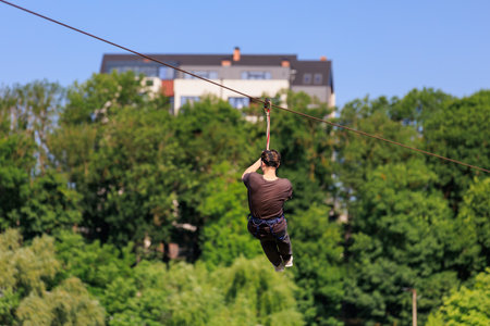 Male adult ziplining through greenery with building in background under blue sky.の写真素材