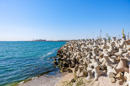Coastal breakwaters near industrial port with cranes and blue sea.の写真素材