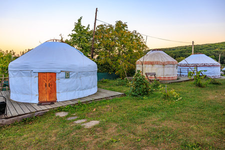 A white tent with a wooden door sits in a grassy field. The tent is surrounded by a fence and a path leading to it. The scene is peaceful and serene, kyrgyz authentic nomadic yurtの写真素材
