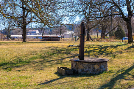 Rustic water well in open field with trees and farm buildings in background.の写真素材