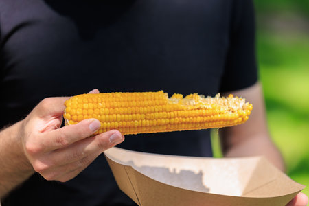 Male holding corn on the cob in sunny park setting.