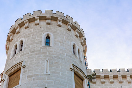 Medieval-style stone castle turret against blue sky with arrow slits and battlements.の写真素材