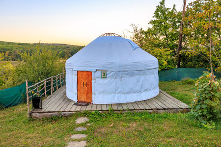 A white tent with a wooden door sits in a grassy field. The tent is surrounded by a fence and a path...