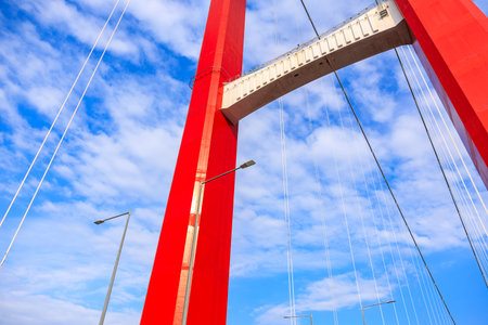 Red suspension bridge towering against blue sky with clouds.の写真素材