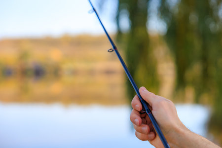 Male hand holding fishing rod near calm lake.