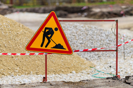 A bright warning sign marks a construction zone with workers digging. Gravel and dirt piles surround...