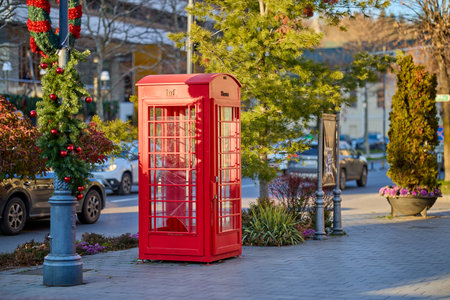 Vintage red telephone booth on leafy street in urban setting. December 13, 2025 Sinaia Romania.のeditorial素材