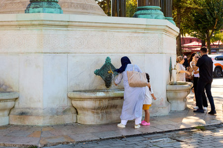 Woman in hijab with child at outdoor fountain in sunny urban park setting. August 10, 2025, Istanbul Turkey.のeditorial素材