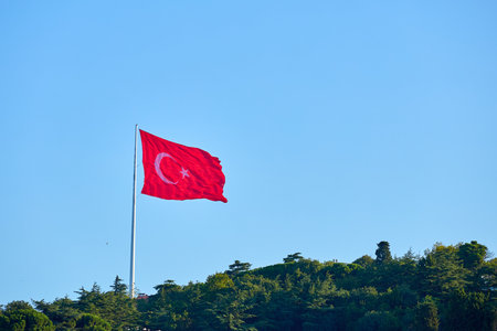 Turkish flag waving on hilltop with clear blue sky and surrounding greenery.