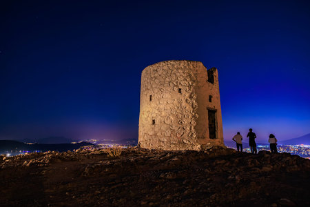Ancient windmill at night with people viewing cityscape under starry sky.の写真素材