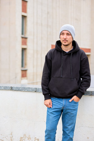 Young man stands casually on urban rooftop. Gray beanie, black hoodie, blue jeans complete his look....