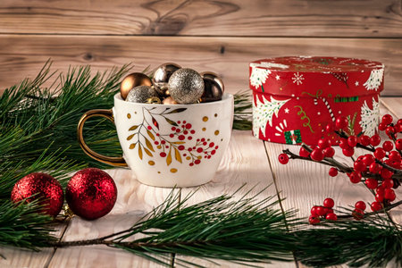 White cup with Christmas balls, natural pine branches and red ilex berries around. Winter still life with gift red box on a light wooden background.の写真素材