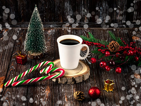 A white cup of coffee and red and green Christmas lollipops in the shape of a heart on a table on a dark background. Christmas tree, balls, branch of holly, cones as decoration.の写真素材
