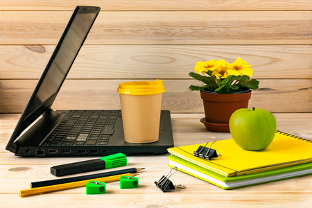 Office workplace with open laptop, notebooks, paper cup of coffee, marker on wooden table. A green apple and a pot with a flower as decor. business background.の写真素材
