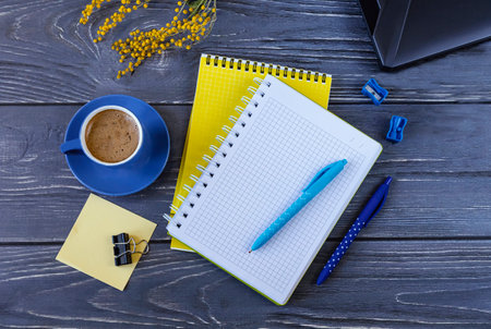 Top view of workplace with notebooks, pens, marker, laptop and blue cup of coffee and dark table. A branch of mimosa as a decor business background.の写真素材