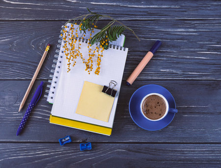 Top view of workplace with notebooks, pens, marker, laptop and blue cup of coffee and dark table. A branch of mimosa as a decor business background.の写真素材