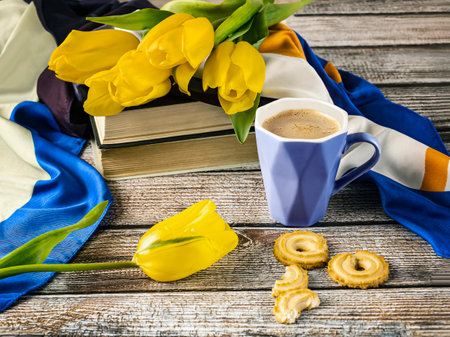A blue cup of coffee and a bouquet of yellow tulips. Spring still life with book, scarf and cookies on wooden background.の写真素材