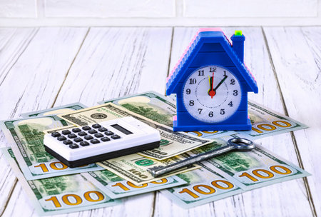 real estate loans. Dollar bills, an alarm clock in the form of a house and an apartment key and a calculator on a white wooden background. Investments in constructionの写真素材