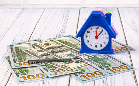 Money for real estate, home loan concept. Dollar banknotes, an alarm clock in the form of a house and a key to an apartment on a white wooden background. Investments in real estateの写真素材