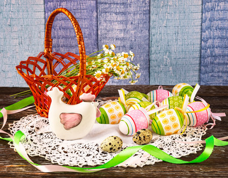 Easter composition with a basket with Easter eggs, decorative chicken, quail eggs on a wooden background. Chamomile flowers and green ribbon as decor.の写真素材