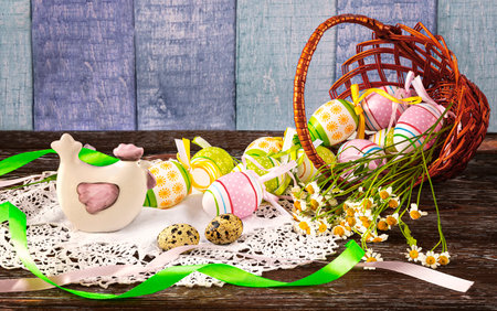 Easter composition with a basket with Easter eggs, decorative chicken, quail eggs on a wooden background. Chamomile flowers and green ribbon as decor.の写真素材
