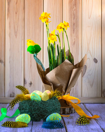 Happy Easter concept. Easter still life with a nest with Easter eggs, yellow daffodil flowers, feathers, decorative chickens, ribbons on a light wooden background.の写真素材