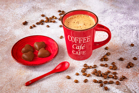 A red cup of coffee with a spoon, coffee beans and chocolate candies in the form of a heart on a light background.の写真素材