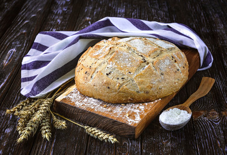 Freshly baked bread with flax seeds, linen towel, wooden tray, wheat ears, wooden spoon with flour on a dark rustic background. front viewの写真素材