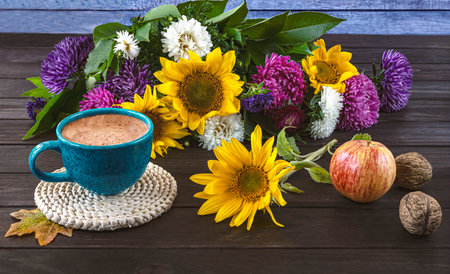 Autumn still life with a bouquet of asters and sunflowers, a blue cup of coffee, an apple and nuts on a dark wooden background.の写真素材